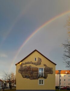 Blick aus der Kirche: Haus mit Lokomotive auf der Fassade und darüber einen doppelten Regenbogen.